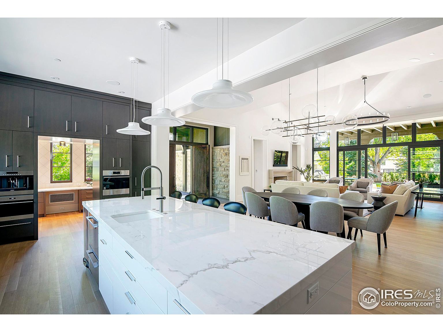 This is a bright, open-concept kitchen and dining area featuring a large white marble island with a sink and modern faucet. Dark gray cabinetry lines one wall, housing built-in appliances, while the dining area boasts a long table with gray chairs and unique pendant lighting. Large windows offer ample natural light and a view of the outdoor space, creating a seamless indoor-outdoor living experience.