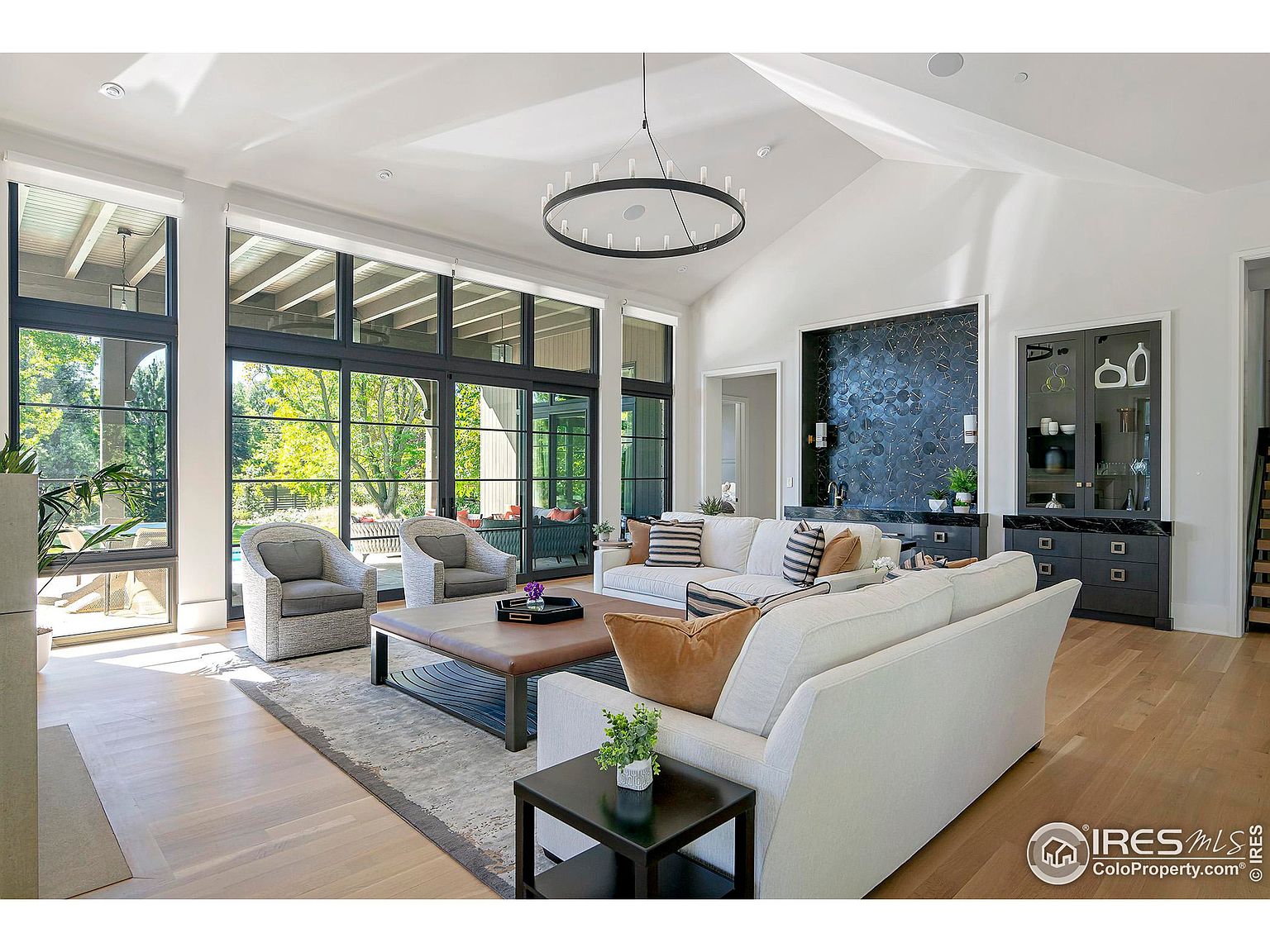 This is a bright and spacious living room featuring large windows that offer ample natural light and views of the outdoors. The room is furnished with comfortable seating, including sofas and armchairs, arranged around a coffee table and a stylish area rug. A modern chandelier hangs from the high ceiling, and a built-in bar area adds a touch of luxury.