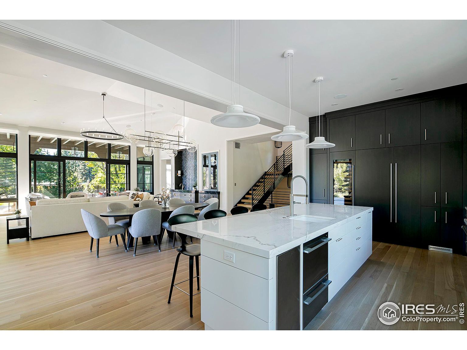 This is a bright and modern kitchen with an open floor plan that includes a dining area and living room. The kitchen features a large island with a white countertop, sleek white cabinetry, and modern pendant lighting. Dark cabinetry lines one wall, housing appliances and providing ample storage, while large windows offer natural light and views of the outdoors.
