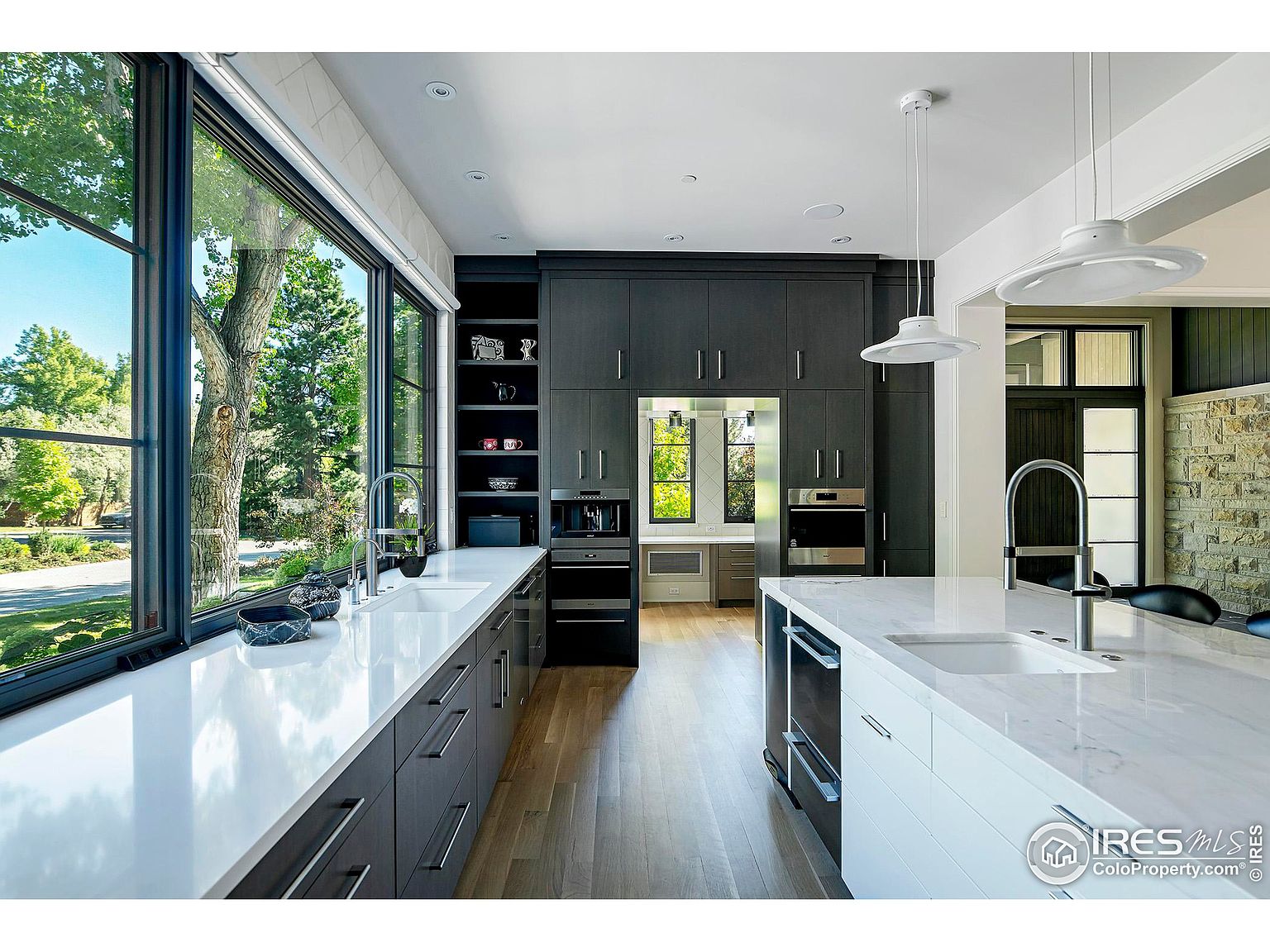 This is a modern kitchen with dark gray cabinetry, white countertops, and stainless steel appliances. A large window provides ample natural light and a view of the outdoors. The kitchen island features a sink and additional storage, and pendant lights hang above it, adding a touch of elegance.