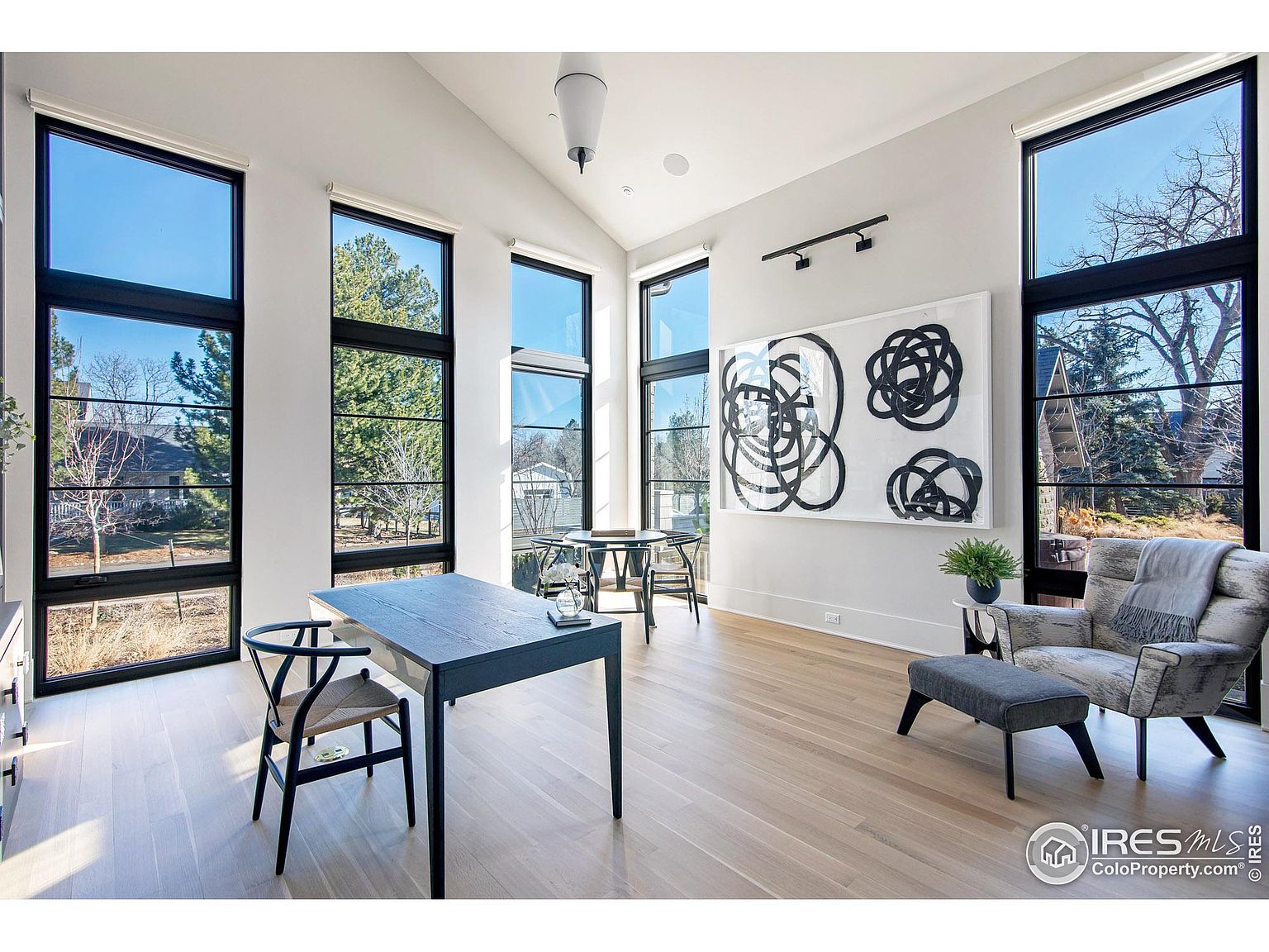 This is a bright and airy home office featuring large windows with black frames that offer views of the outdoors. The room is furnished with a dark-colored desk, a modern chair, and a comfortable armchair with an ottoman. A large piece of abstract art hangs on the wall, adding a touch of sophistication to the space. The light wood flooring and white walls create a clean and inviting atmosphere.
