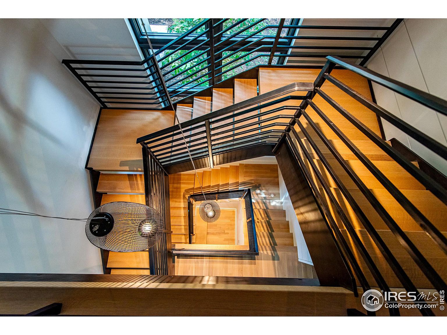 This is a high-angle shot looking down a modern staircase. The stairs are made of light-colored wood with black metal railings. A unique, spherical light fixture hangs in the stairwell, and natural light streams in from above, creating a bright and airy feel.