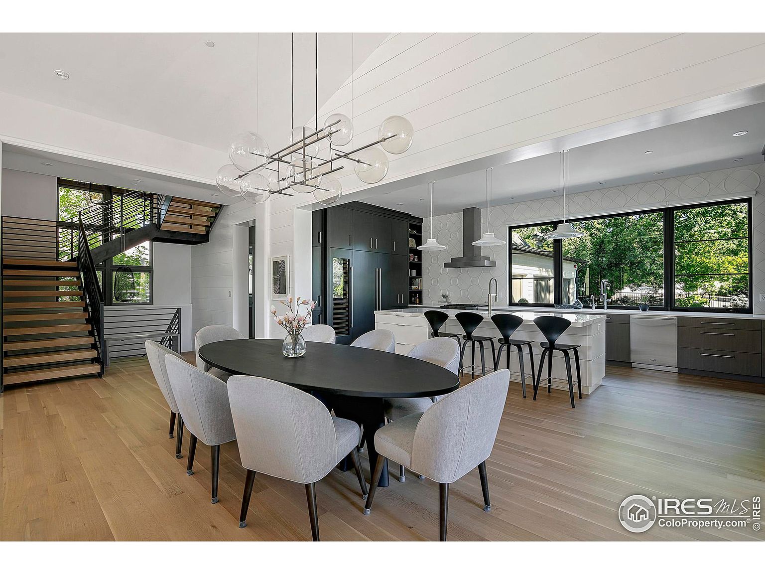 This interior shot showcases a modern dining room adjacent to a kitchen area. The dining room features a dark oval table surrounded by eight light gray upholstered chairs, illuminated by a contemporary chandelier with glass orbs. A staircase is visible to the left, and the kitchen boasts a large window overlooking greenery, creating a bright and inviting atmosphere.