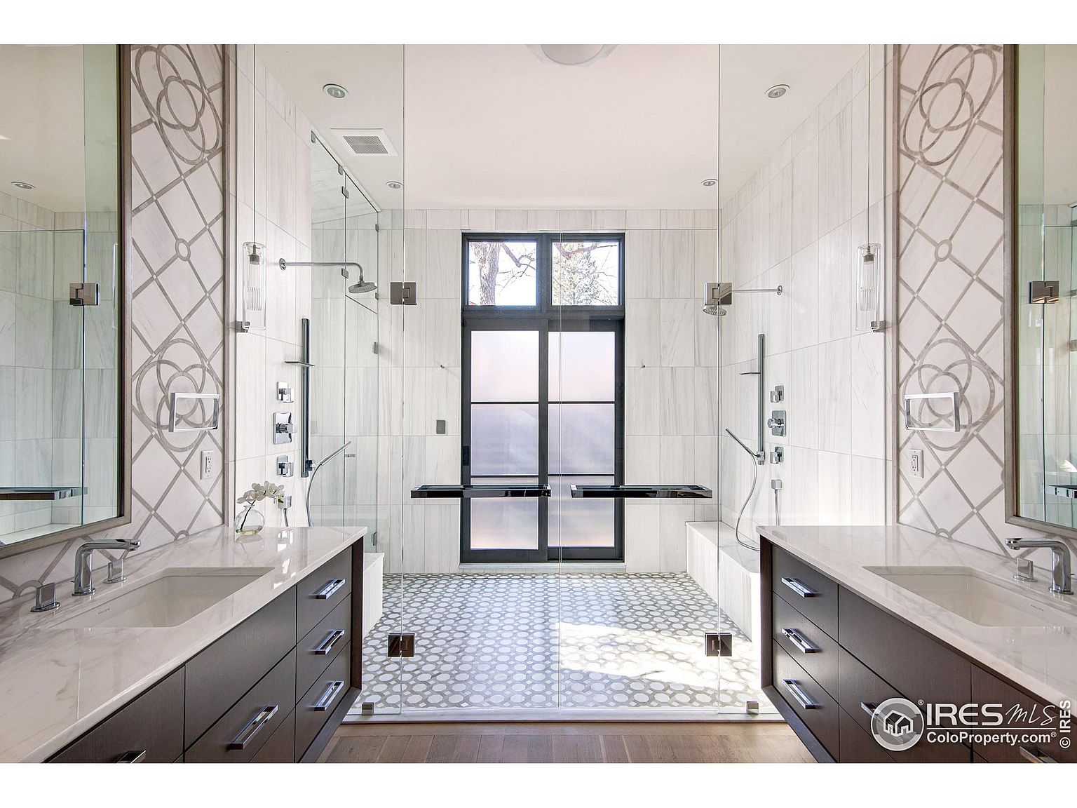 This is a luxurious primary bathroom featuring dual vanities with dark wood cabinets and marble countertops. A large, frameless glass shower enclosure separates the vanities, showcasing a modern shower system and patterned tile flooring. A window provides natural light, enhancing the spa-like atmosphere of the space.