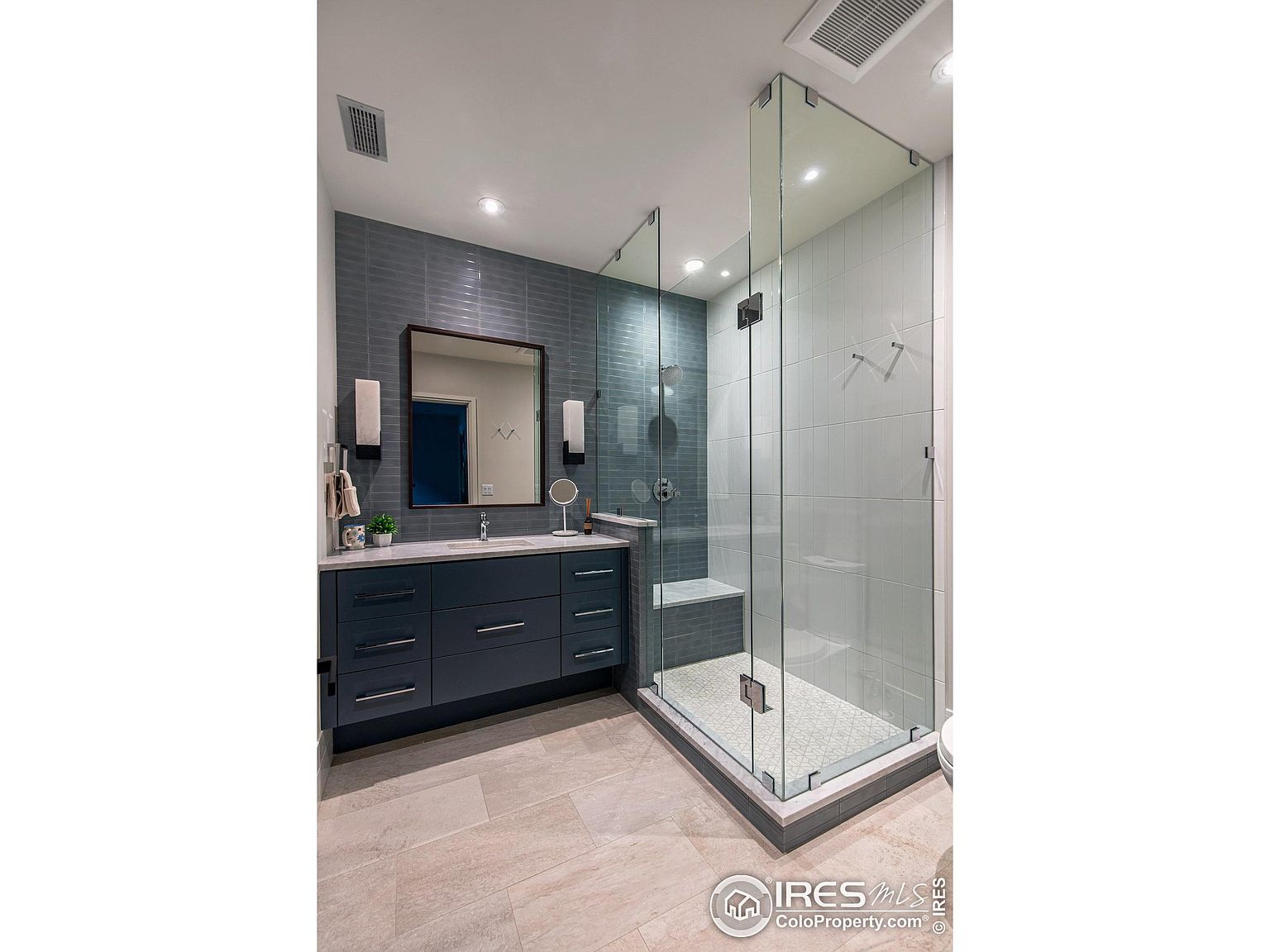 This is a well-lit primary bathroom featuring a modern design. The bathroom includes a vanity with a dark blue cabinet, a white countertop, and a rectangular mirror. A glass-enclosed shower with light-colored tiles and a built-in bench is also visible, creating a clean and luxurious space.