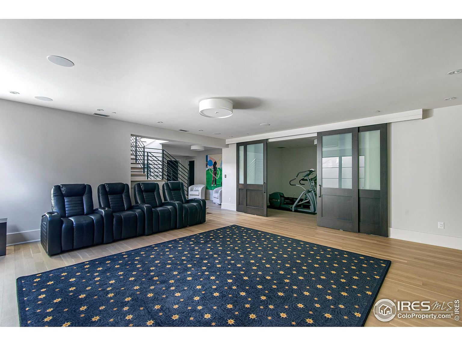 The image showcases a modern living room with a home theater setup, featuring a row of black leather recliners facing the camera. A navy blue rug with gold star patterns covers the light hardwood floor, adding a touch of elegance. Sliding glass doors lead to a home gym, enhancing the room's versatility and appeal.