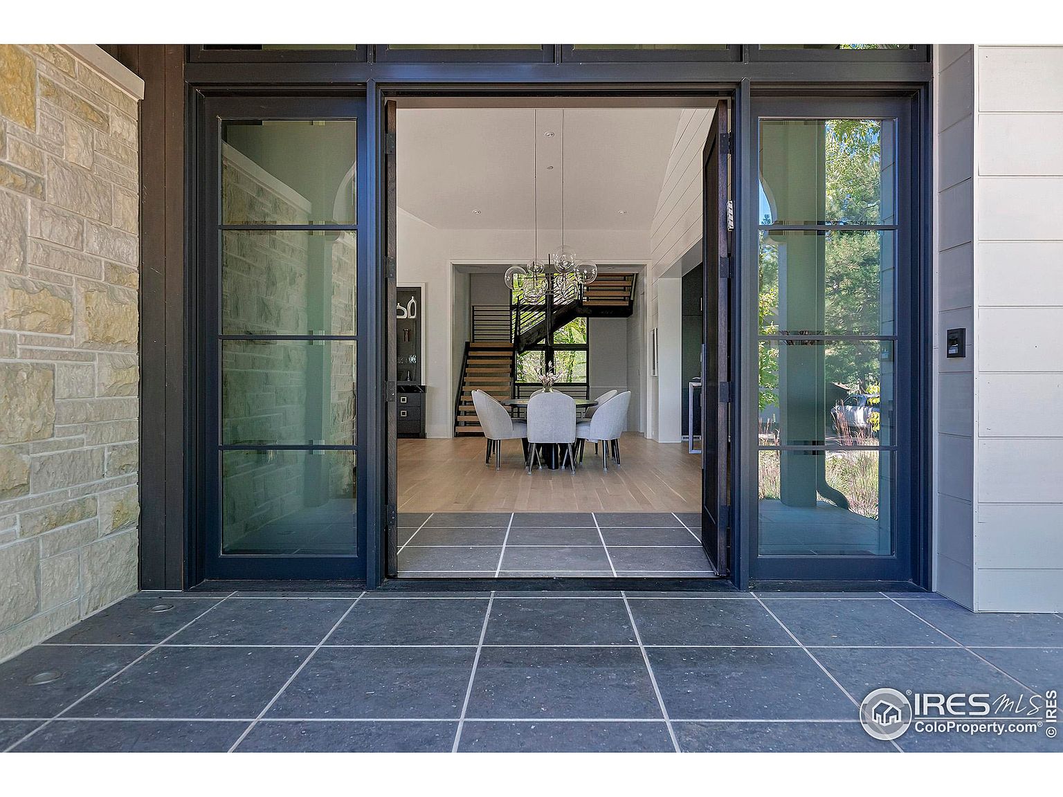 The image showcases an inviting entryway with large, dark-framed glass doors that are partially open, offering a glimpse into a modern dining area with a staircase in the background. The exterior features a combination of stone cladding on one side and light-colored siding on the other, while the entryway is paved with dark, square tiles. The open doors create a seamless transition between the outdoor and indoor spaces, highlighting the home's contemporary design.