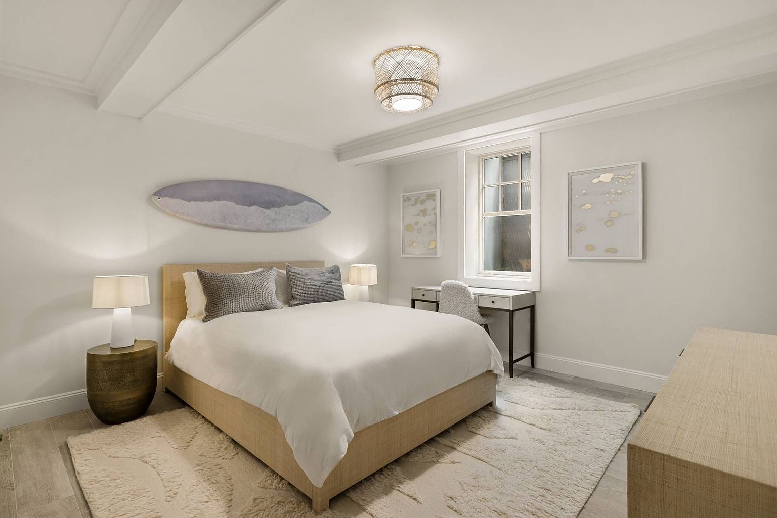 This serene bedroom features a neutral color palette with a plush, textured area rug and a light-toned upholstered bed frame. A surfboard-inspired wall art piece adds a coastal touch above the bed, while a small desk and chair are positioned near the window to create a functional workspace. The room is illuminated by a decorative woven drum pendant light and matching bedside lamps, creating a warm and inviting atmosphere.