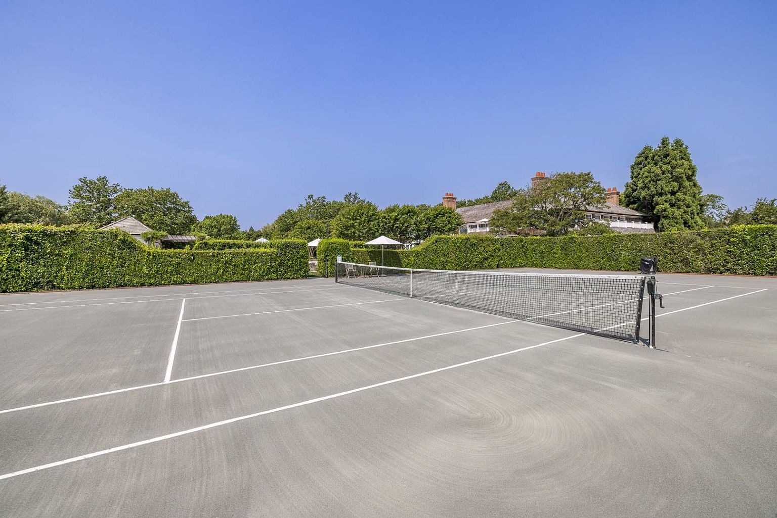 This image features a well-maintained private tennis court set against a backdrop of lush green hedges and mature trees. The court is framed by a clear blue sky, with a glimpse of a traditional-style residence visible in the distance. The perspective is from the court level, emphasizing the expansive playing surface and the quiet, secluded atmosphere of the property.