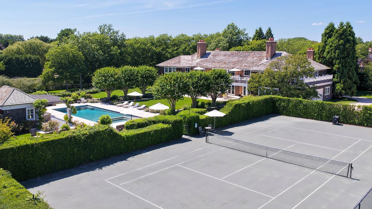 This elevated aerial perspective captures a sprawling estate featuring a large shingle-style residence surrounded by lush greenery. The property includes a private tennis court in the foreground, a swimming pool with an adjacent patio, and meticulously manicured hedges that define the outdoor living spaces. The scene conveys a sense of luxury, privacy, and expansive recreational amenities.