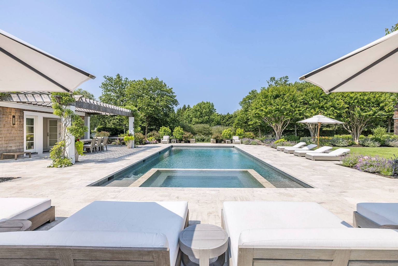 This serene outdoor living space features a rectangular swimming pool with an integrated spa, surrounded by light-toned stone decking. The area is framed by lush green trees and manicured landscaping, with multiple lounge chairs and umbrellas providing comfortable seating. A pergola-covered patio area to the left offers an additional shaded space, creating a luxurious and inviting atmosphere for relaxation.