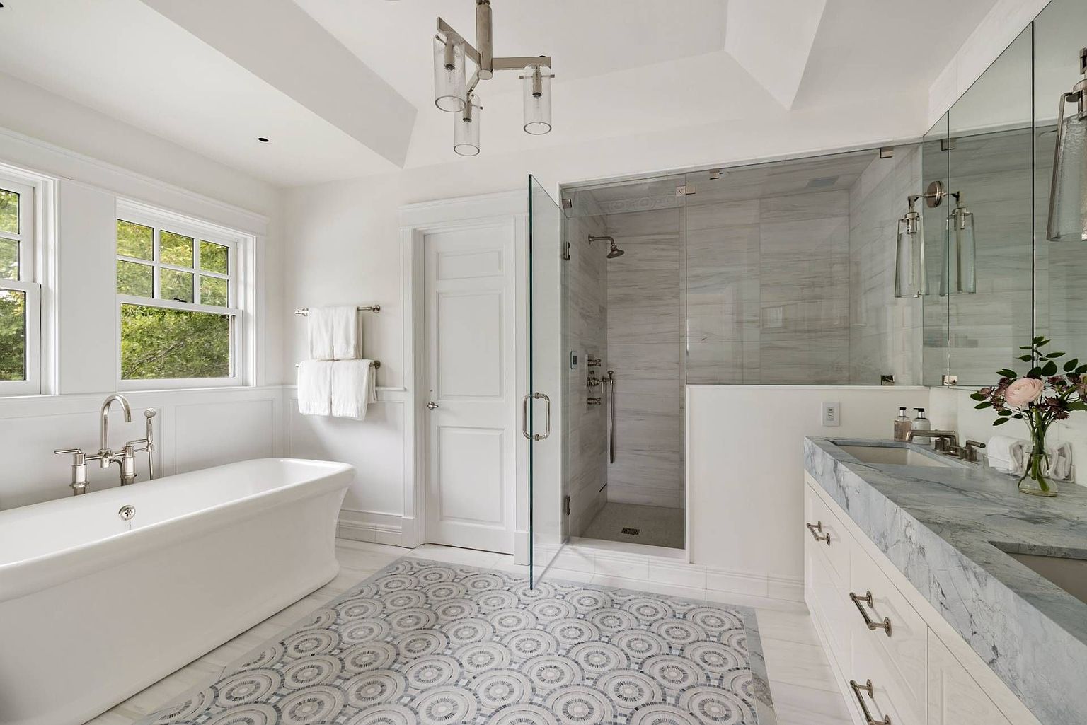 This bright and elegant primary bathroom features a freestanding soaking tub positioned beneath large windows, complemented by a spacious walk-in shower with glass paneling and light-toned stone wall tiles. A double vanity with a stone countertop and polished hardware sits opposite the shower, while a decorative patterned tile rug adds visual interest to the floor. The space is illuminated by a modern chandelier, creating a clean, sophisticated, and airy atmosphere.