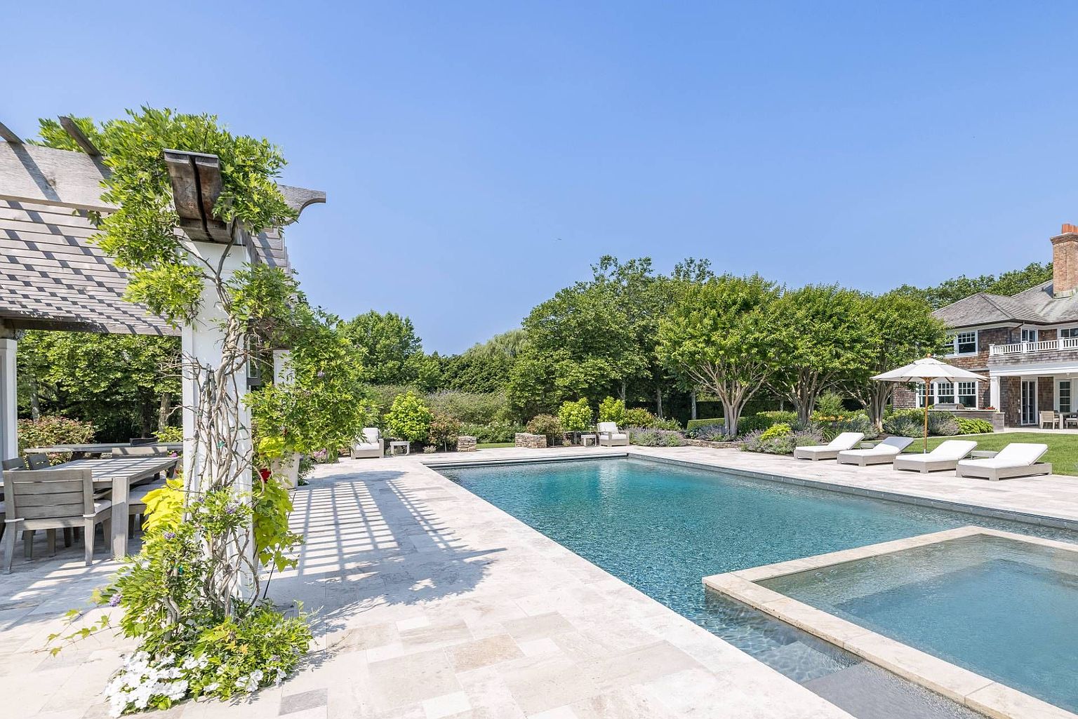 This expansive outdoor living space features a large rectangular swimming pool with an integrated spa, surrounded by light-toned stone decking. To the left, a wooden pergola provides shade for an outdoor dining area, while a row of lounge chairs sits poolside under a white umbrella. The scene is framed by lush green trees and manicured landscaping, with a multi-story shingled house visible in the background, creating a serene and luxurious backyard retreat.