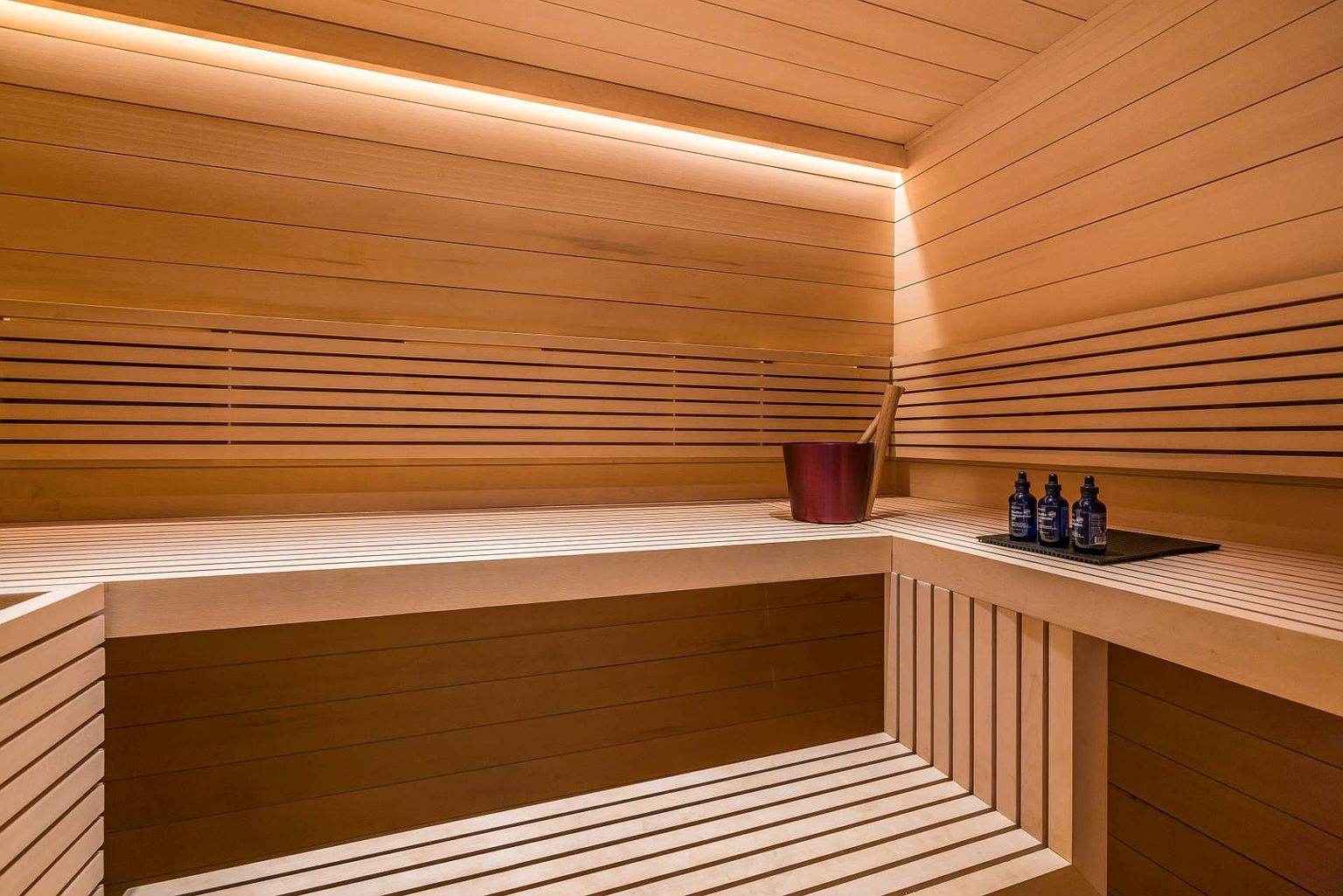 This image showcases a minimalist sauna interior featuring light-toned wood paneling and tiered bench seating. A metallic bucket and wooden ladle sit on the upper bench, accompanied by a small tray of essential oil bottles, creating a serene and spa-like atmosphere. The space is illuminated by a warm, recessed linear light strip that highlights the clean lines and natural texture of the wood.