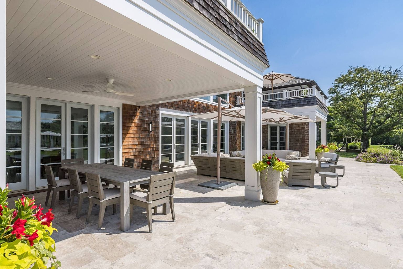 This expansive stone-paved patio offers a luxurious outdoor living space, featuring a covered dining area with a wooden table and chairs, alongside a comfortable lounge section shaded by large umbrellas. The home showcases classic shingle-style architecture with multiple glass doors providing seamless indoor-outdoor flow. The scene is bathed in bright, natural sunlight, highlighting the well-maintained landscaping and the serene, upscale atmosphere of the backyard.
