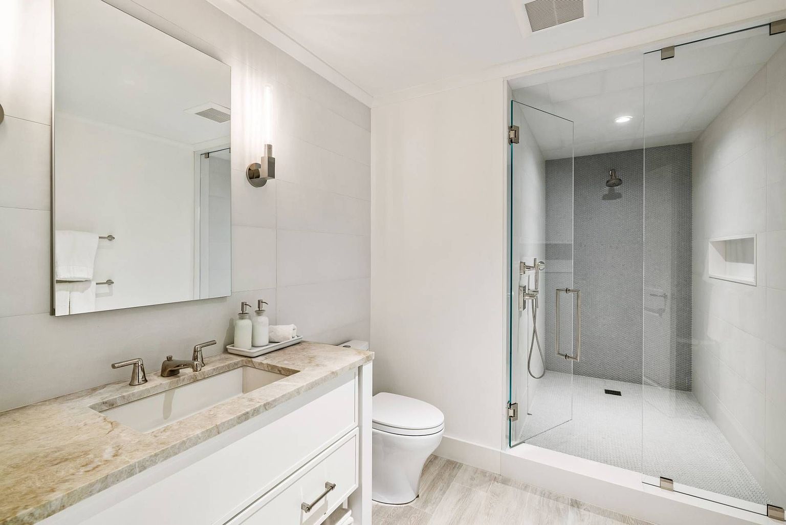 This modern bathroom features a sleek vanity with a stone countertop and a rectangular undermount sink, paired with a large, frameless mirror. To the right, a spacious walk-in shower is enclosed by glass panels and showcases a textured, dark-toned tile accent wall with a built-in niche. The room is finished with light-colored wall tiles and wood-look flooring, creating a bright and contemporary aesthetic.