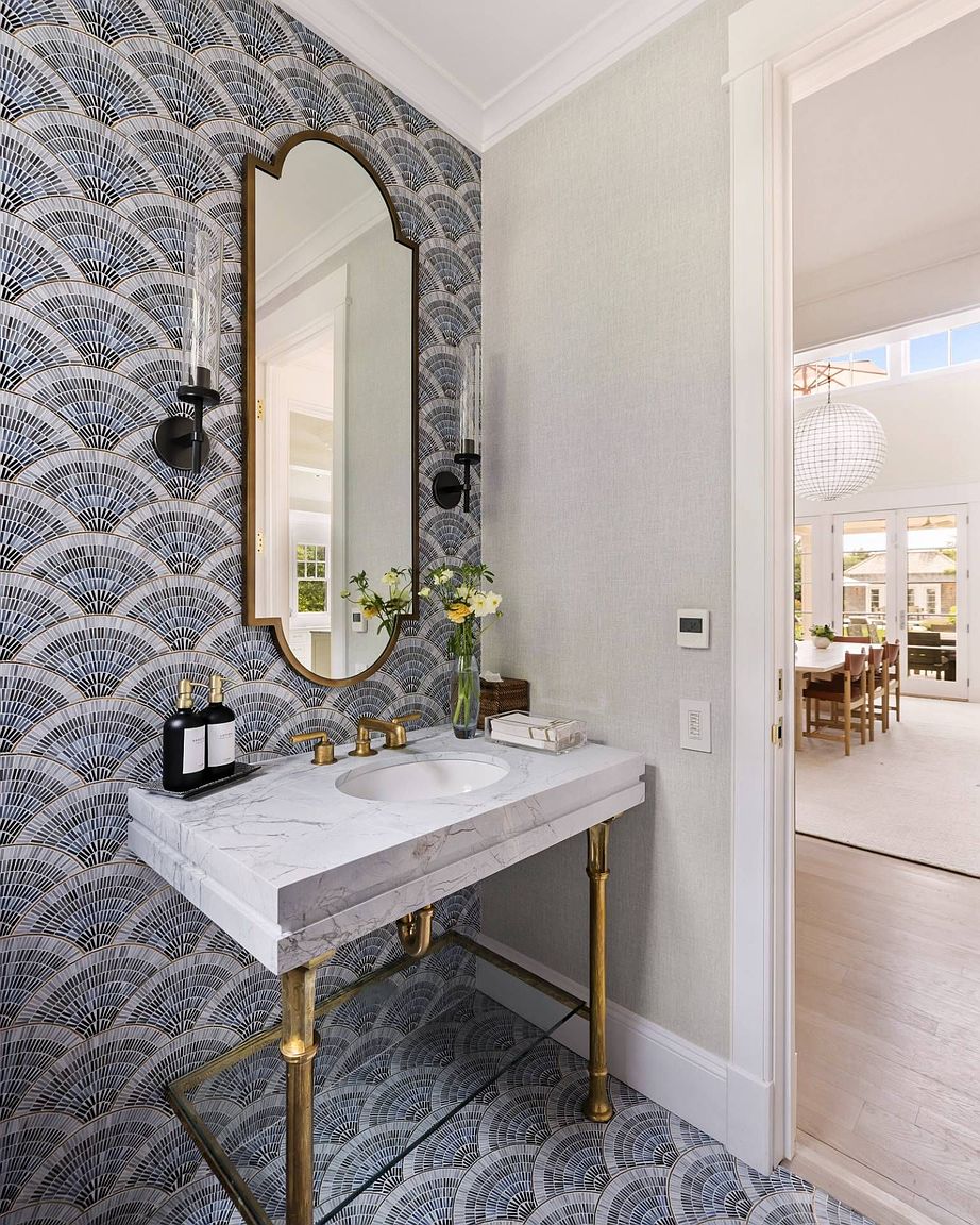 This elegant powder room features striking fan-patterned wallpaper that extends from the walls to the floor, creating a cohesive and bold aesthetic. A stone-topped vanity with a brass-toned metal base is centered beneath a decorative arched mirror, flanked by two wall-mounted light fixtures. Through the open doorway, a glimpse of a bright, airy dining area with a large pendant light provides a sense of depth and connection to the rest of the home.