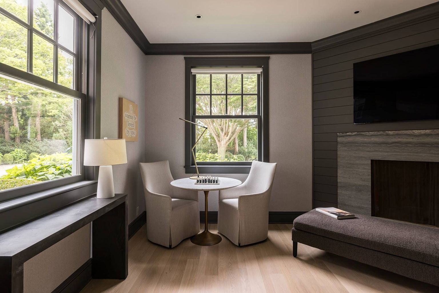This sophisticated living space features a cozy seating area with two upholstered chairs flanking a small round table, positioned perfectly by a window overlooking lush greenery. To the right, a dark-toned wall with horizontal paneling frames a television mounted above a stone-clad fireplace surround, while a long bench provides additional seating. The room is finished with dark trim, light wood flooring, and a minimalist table lamp, creating an inviting and refined atmosphere.