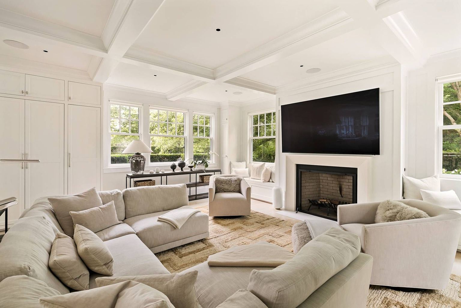 This bright and airy living room features a large, neutral-toned sectional sofa and two matching armchairs arranged around a textured area rug. A television is mounted above a fireplace with a clean, light-colored surround, while built-in cabinetry and window seats provide ample storage and cozy seating. The space is bathed in natural light from multiple windows, creating a sophisticated and inviting atmosphere.