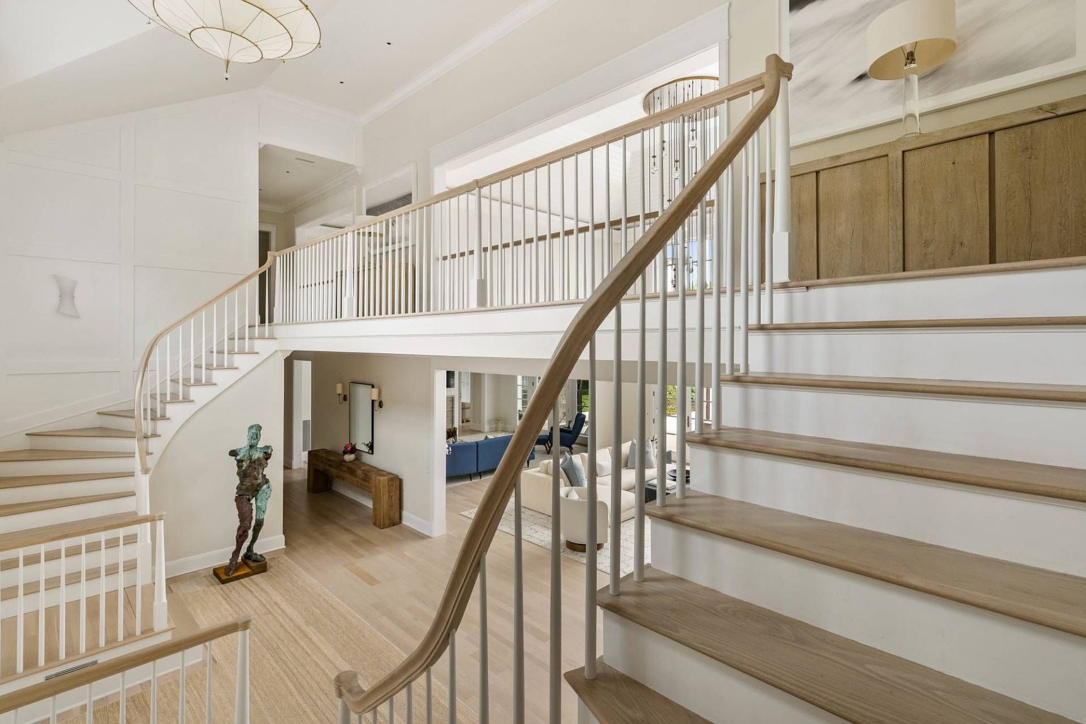 This grand foyer features a sweeping, curved staircase with light wood treads and white balusters that serves as the architectural centerpiece of the home. The open-concept design provides a clear view into the living area below, while a sculptural art piece stands prominently on the lower level. The space is characterized by bright, neutral tones, clean lines, and a sophisticated, airy atmosphere.