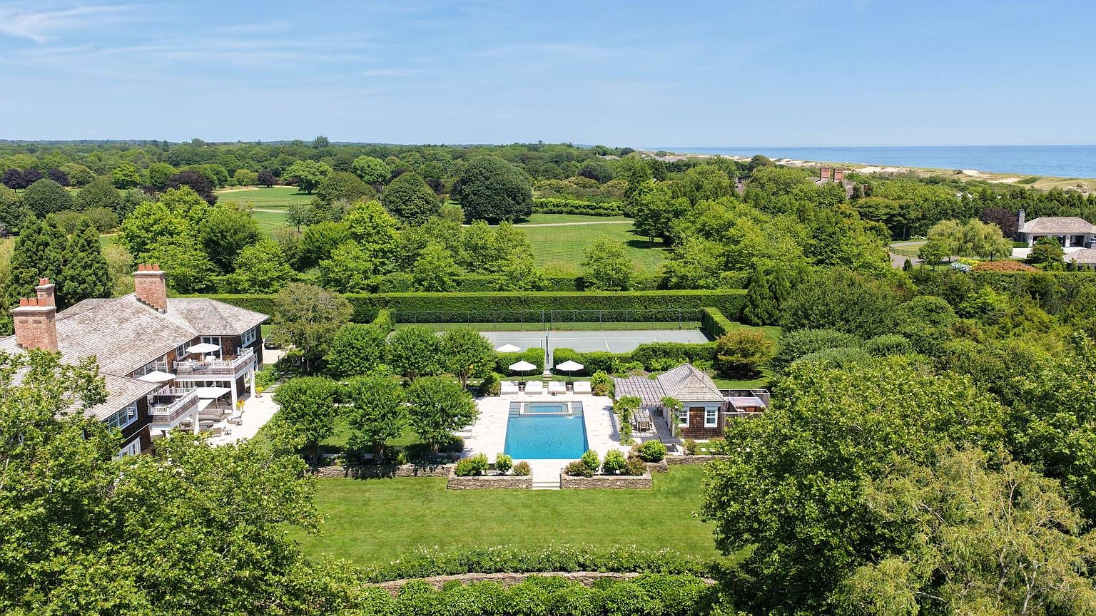 This expansive aerial view captures a luxurious coastal estate featuring a shingle-style main residence, a private swimming pool with an adjacent pool house, and a tennis court. The property is surrounded by lush, manicured landscaping and mature trees, with a vast green lawn extending toward the distant ocean horizon. The scene conveys a sense of privacy and high-end leisure in a serene, sun-drenched setting.