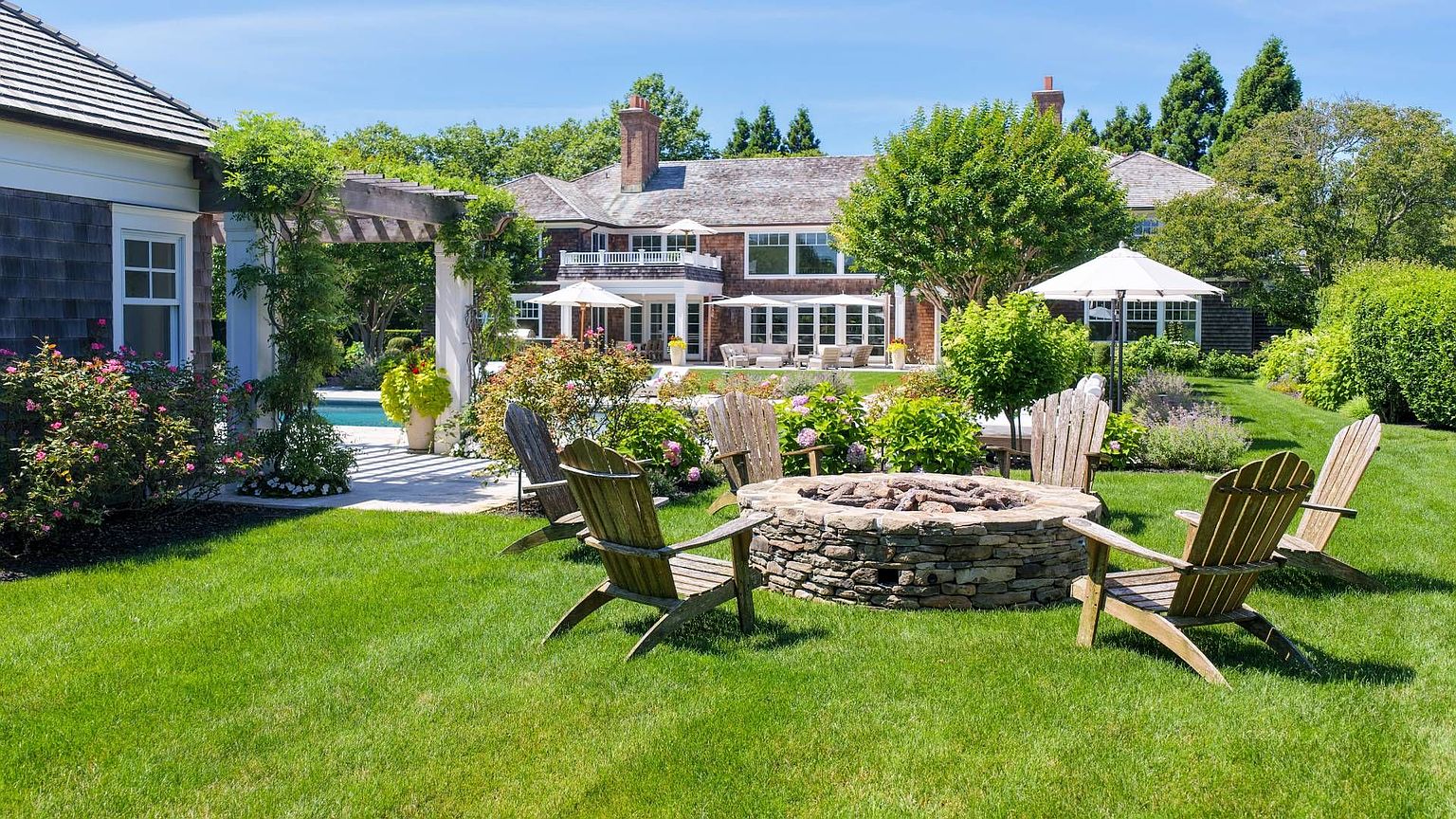This expansive backyard features a lush green lawn centered around a circular stone fire pit surrounded by Adirondack chairs. A wooden pergola covers a stone patio area near the house, which showcases classic shingle-style architecture with multiple windows and a second-story balcony. The scene is framed by mature trees and manicured landscaping, creating a serene and inviting outdoor living space.