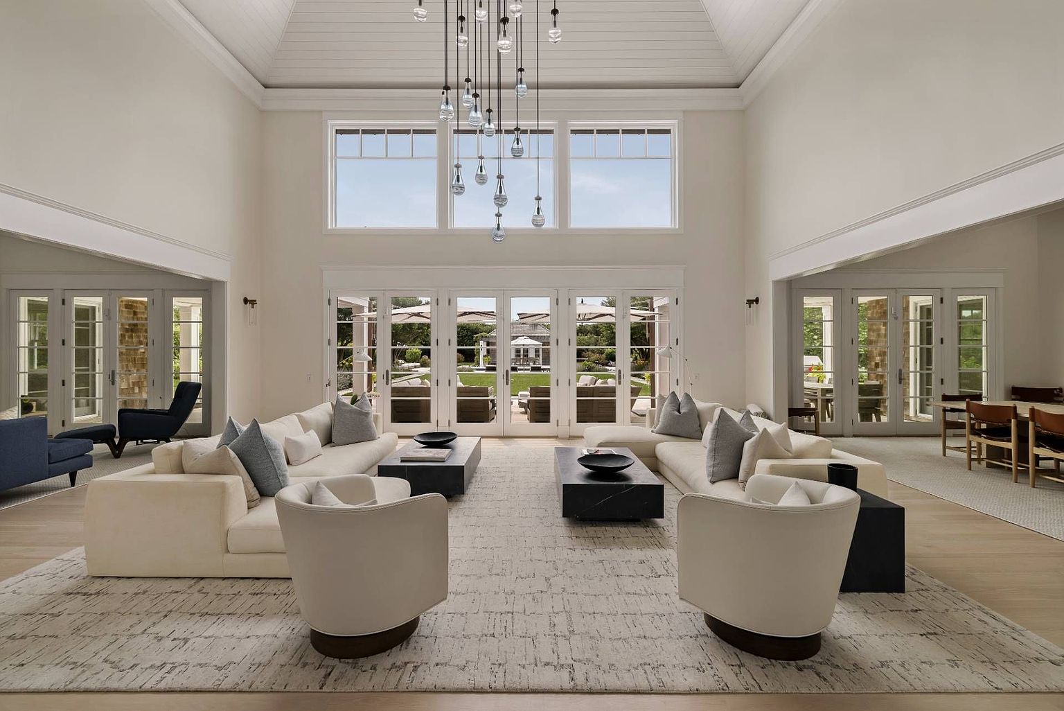 This expansive, high-ceilinged living room features a symmetrical layout with two large white sofas facing each other across dark, low-profile coffee tables. A cluster of modern pendant lights hangs from the vaulted ceiling, drawing the eye toward the large windows that offer a clear view of the backyard. The space is anchored by a textured area rug and two curved accent chairs, creating a sophisticated and airy atmosphere perfect for entertaining.