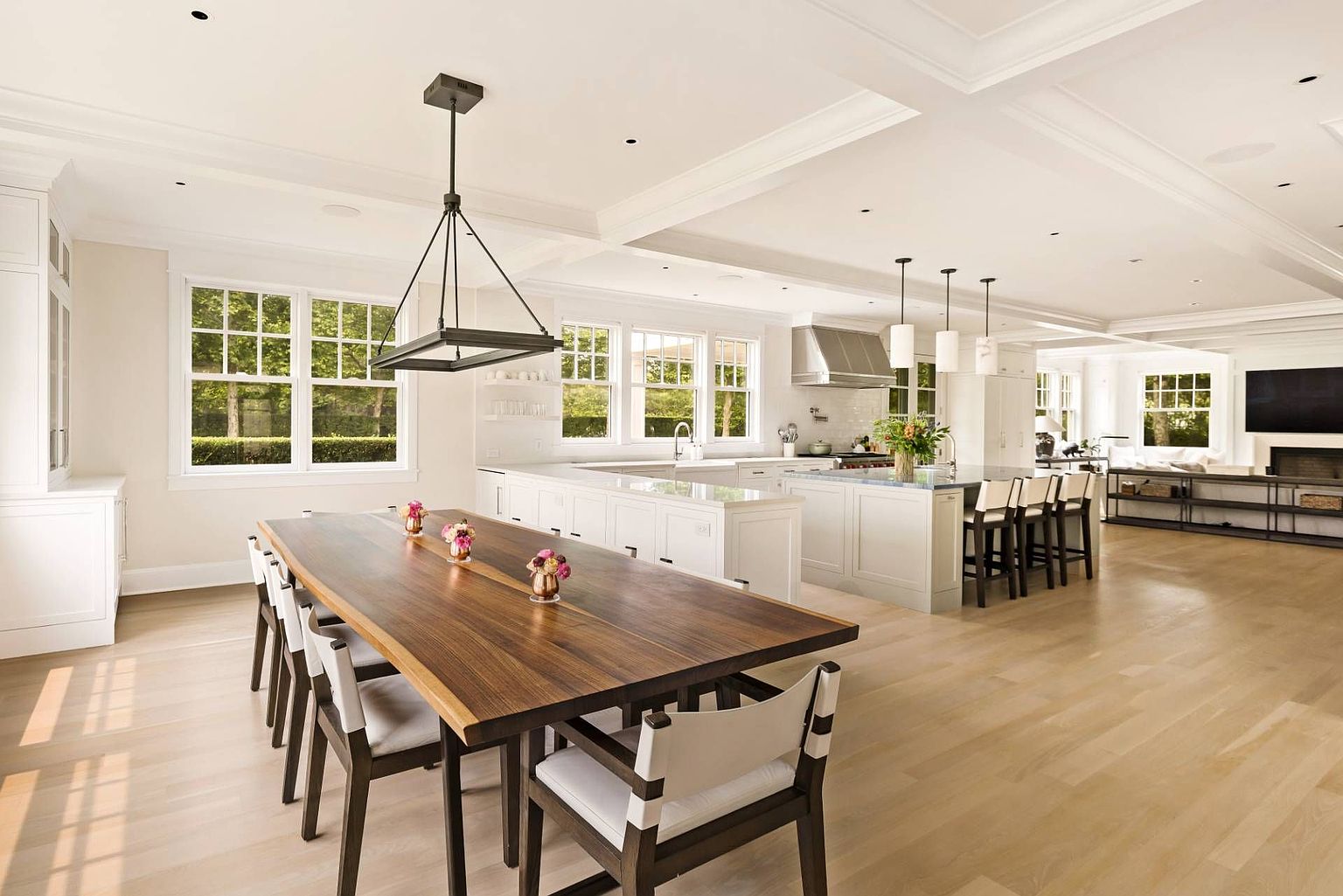 This open-concept space seamlessly blends a dining area with a large, modern kitchen. A long wooden dining table sits in the foreground under a geometric pendant light, while the kitchen features a spacious island with bar seating, white cabinetry, and light wood flooring throughout. The perspective captures the airy, interconnected layout that flows into a living area with a television and fireplace in the background.