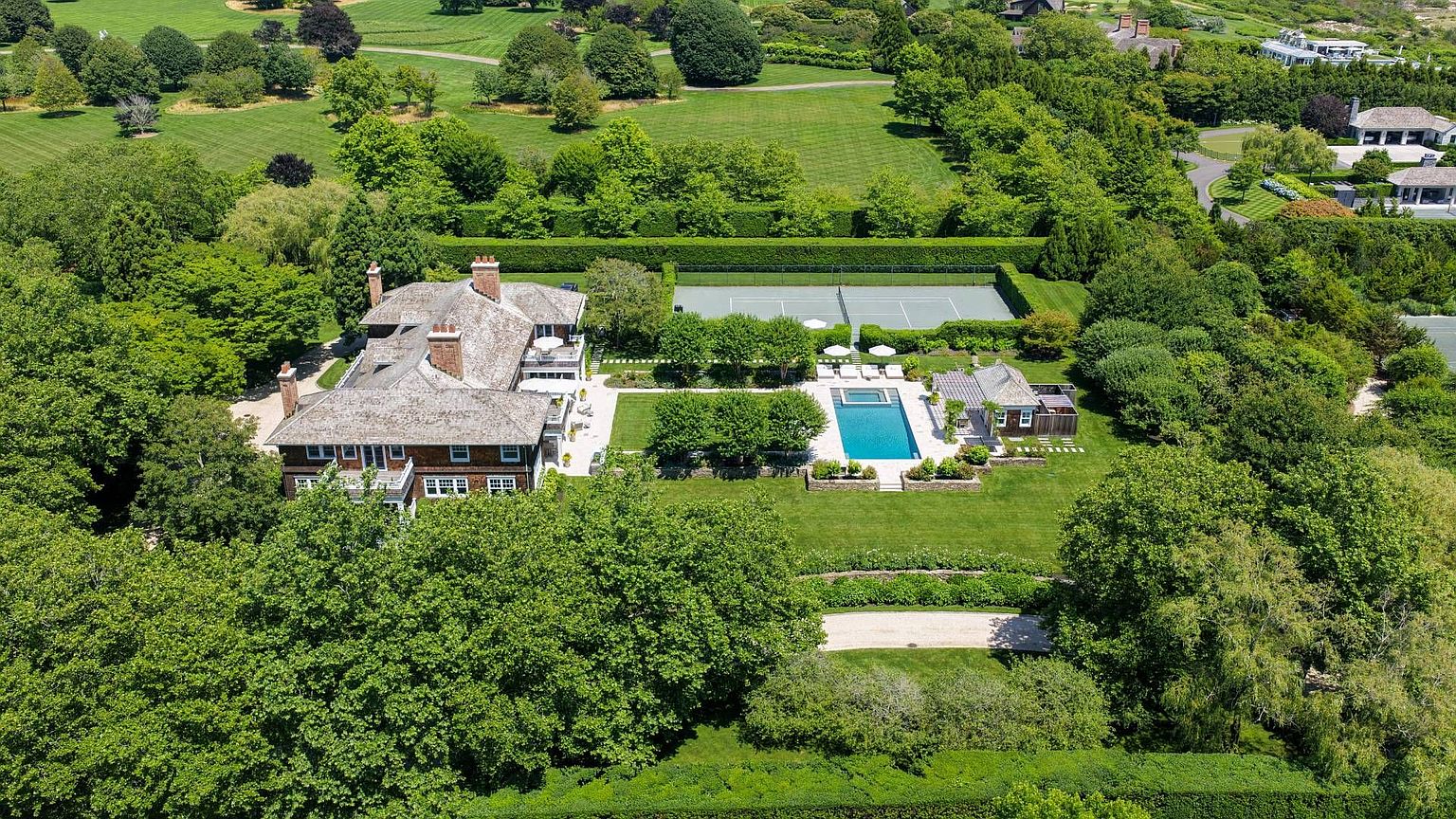 This high-angle aerial view captures a sprawling estate featuring a large, multi-gabled shingle-style residence nestled within lush, mature landscaping. The property includes a private tennis court, a rectangular swimming pool with an adjacent pool house, and expansive manicured lawns surrounded by dense trees. The perspective provides a comprehensive look at the layout of the grounds and the integration of the home into its serene, park-like setting.