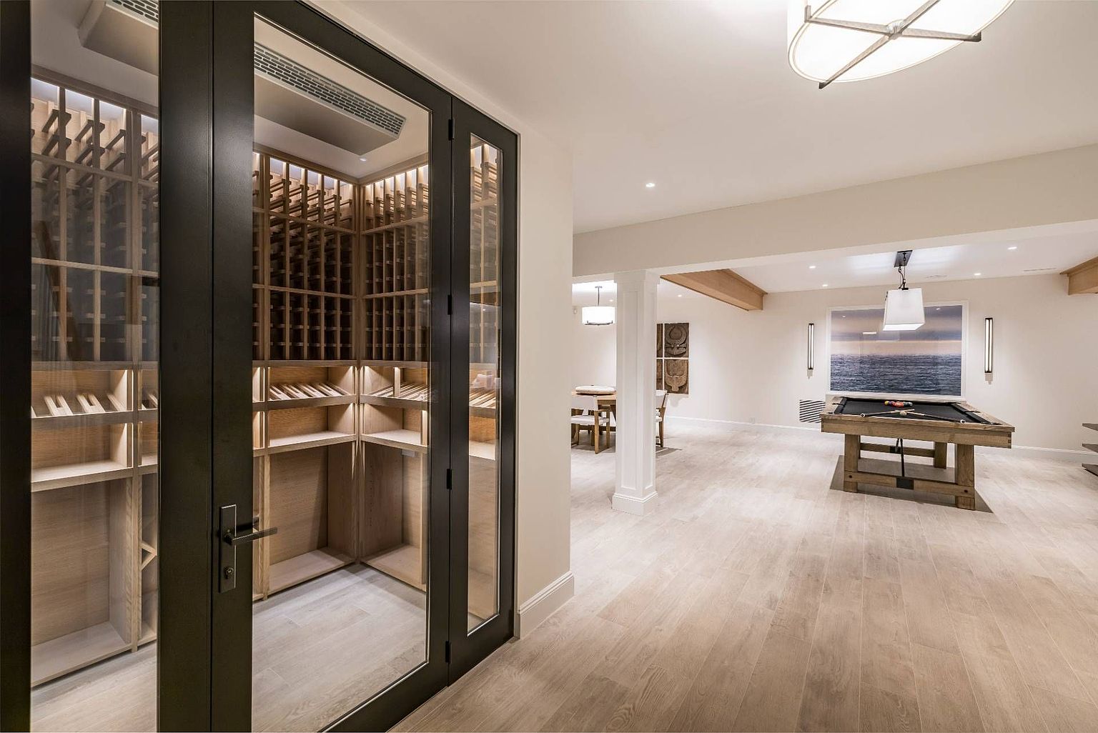 This spacious basement features a modern wine cellar enclosed by glass and dark metal framing, positioned adjacent to a large recreation area. The open-concept space includes a pool table centered under a pendant light, light-toned wood flooring, and a neutral color palette throughout. The perspective looks from the wine cellar entrance across the room, highlighting the clean, contemporary design and versatile layout.
