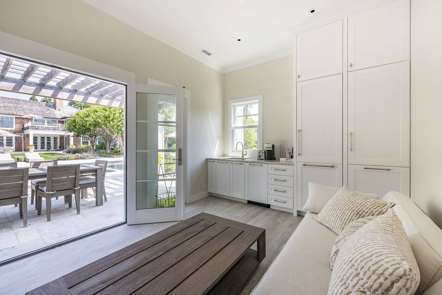 This bright and airy interior space functions as a versatile living area, featuring a light-toned sofa and a low-profile wooden coffee table. A kitchenette with white cabinetry and a sink is tucked along the wall, while large glass doors open directly onto a sunny patio with outdoor dining furniture. The room is bathed in natural light, creating a seamless transition between the indoor living space and the outdoor pool area.