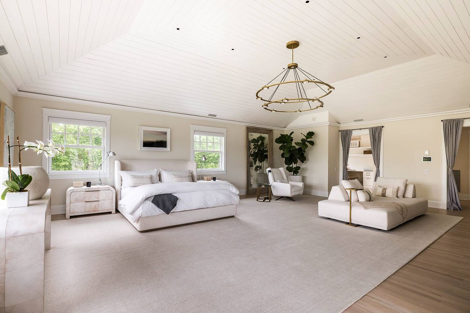 This expansive primary bedroom features a neutral color palette, vaulted ceilings with white paneling, and a large chandelier as a central focal point. The room is furnished with a plush bed, a modern lounge area, and a tall mirror, all set against a backdrop of soft carpeting and light-toned walls. Large windows allow ample natural light to fill the space, creating an airy and sophisticated atmosphere.