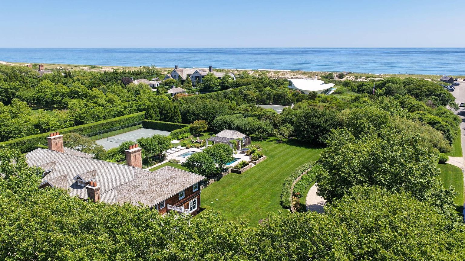 This high-angle aerial view captures a sprawling coastal estate nestled among lush greenery, featuring a large shingle-style residence with multiple brick chimneys. The property includes a private tennis court, a swimming pool area with a patio, and expansive manicured lawns that lead toward the ocean. The perspective highlights the home's secluded setting, its proximity to the beach, and the surrounding natural landscape.