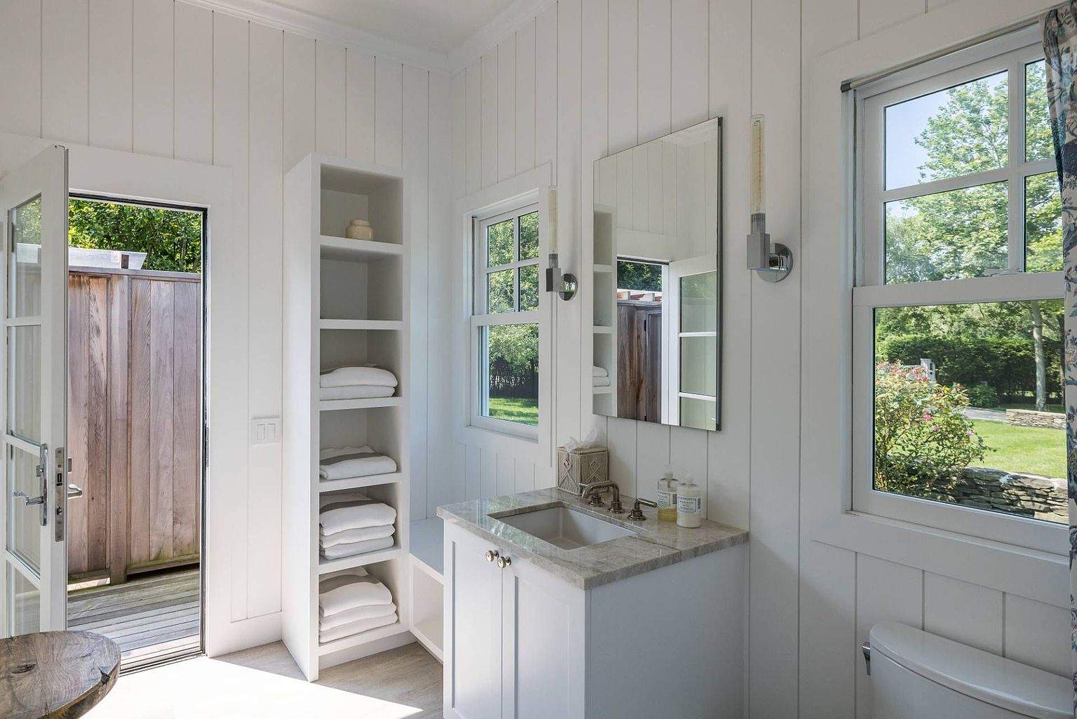 This bright, coastal-inspired bathroom features white vertical shiplap walls and a vanity with a stone countertop. A tall, built-in shelving unit provides organized storage for towels, while a glass door opens directly to an outdoor deck area. The space is filled with natural light from multiple windows, creating an airy and refreshing atmosphere.
