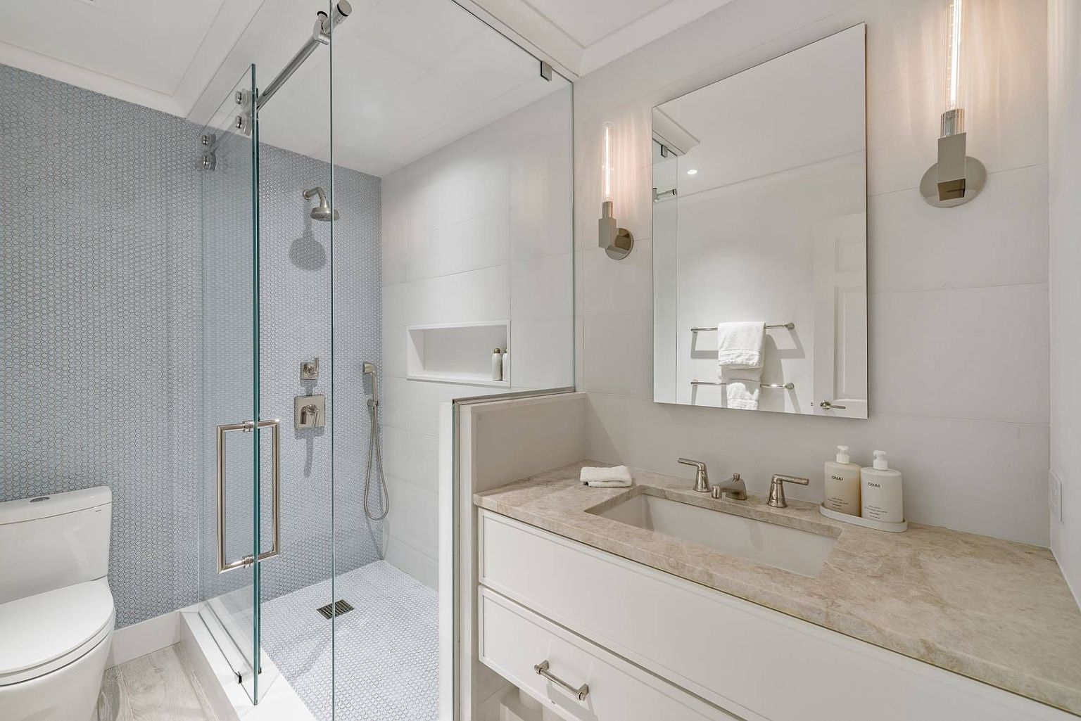 This modern bathroom features a glass-enclosed walk-in shower with patterned tile walls and a matching tiled floor. Adjacent to the shower is a vanity with a stone countertop, a rectangular undermount sink, and a large mirror flanked by two vertical wall sconces. The space is bright and clean, utilizing a neutral color palette to create a contemporary and functional aesthetic.