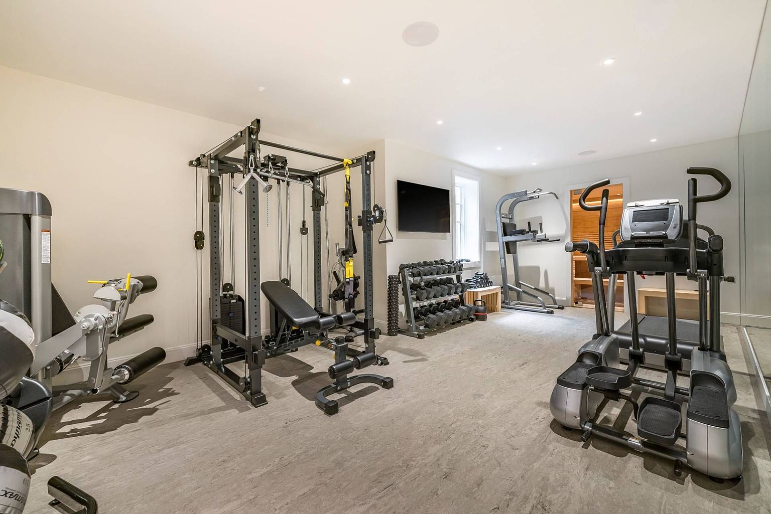 This well-equipped home gym features a variety of professional-grade fitness equipment, including a multi-station cable machine, a rack of dumbbells, and an elliptical trainer. The space is finished with neutral walls, recessed lighting, and durable flooring, creating a functional and dedicated workout environment. A wall-mounted television and a mirrored wall enhance the utility and spacious feel of the room.
