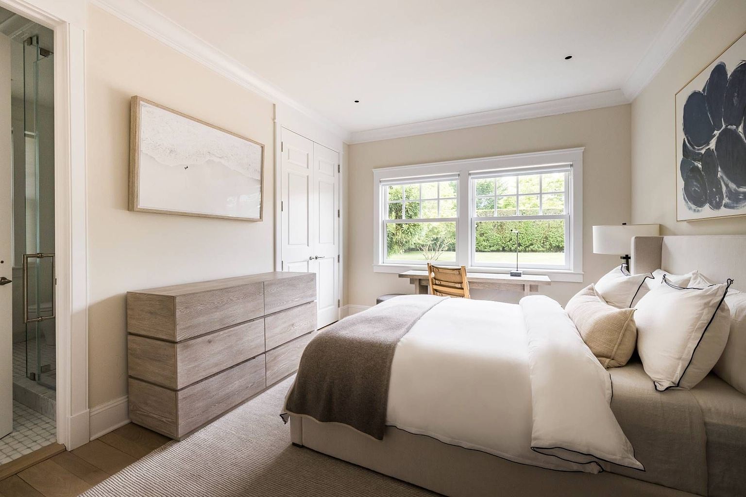 This serene bedroom features a neutral color palette with light wood furniture, including a dresser and a desk positioned by the window. The room is anchored by a plush bed with white linens and a dark throw, complemented by soft wall art and a large window that brings in natural light. The space offers a clean, minimalist aesthetic with a view into an adjacent glass-enclosed shower area.