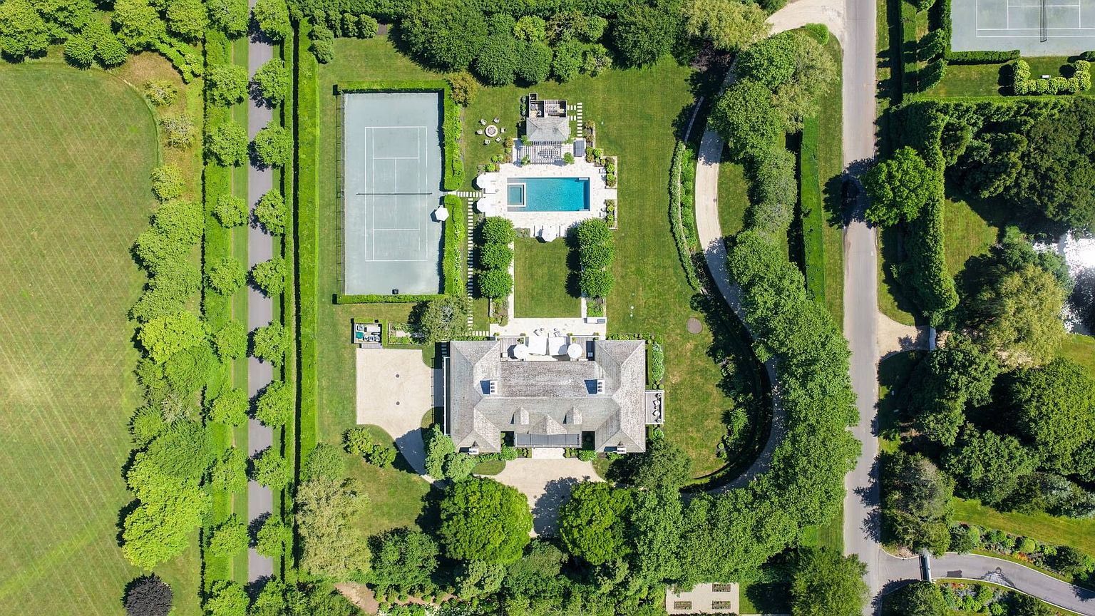 This high-altitude aerial view captures a sprawling estate featuring a large, multi-gabled residence surrounded by lush, manicured landscaping. The property includes a private tennis court, a rectangular swimming pool with an adjacent patio, and extensive tree-lined driveways. The perspective provides a comprehensive look at the expansive grounds, highlighting the symmetry and formal layout of the estate.
