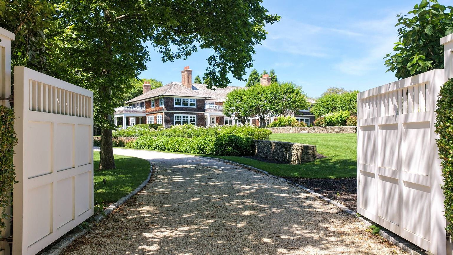 This image captures the grand entrance to a sprawling estate, framed by open white wooden gates that lead onto a winding gravel driveway. The path curves toward a large, multi-story shingled residence nestled among mature trees and lush, manicured landscaping. The scene conveys a sense of privacy and classic elegance, with a stone wall bordering the expansive green lawn.