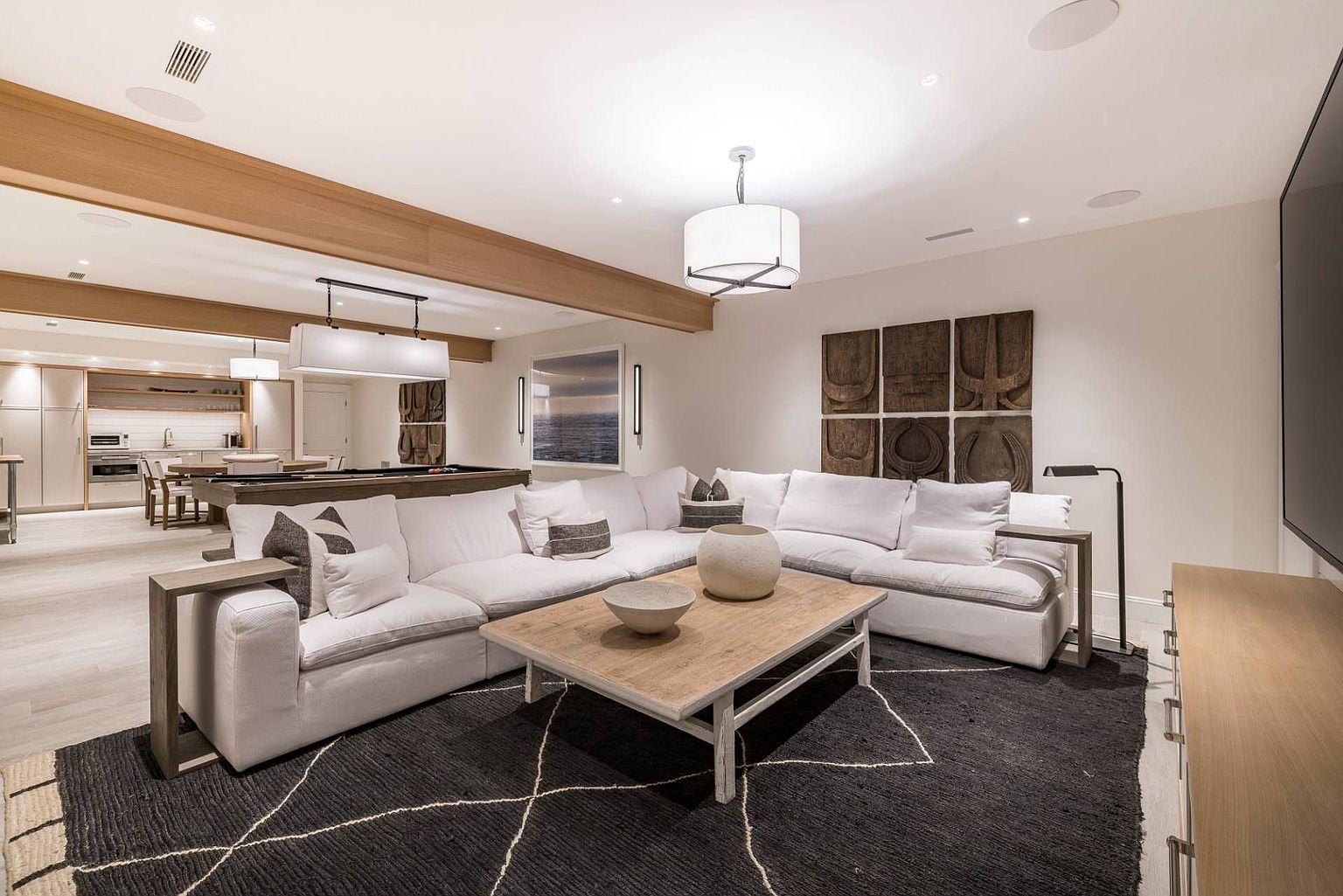 This spacious, open-concept living area features a large white sectional sofa arranged on a dark, patterned area rug, centered around a light wood coffee table. The room is accented by decorative wall art and warm wood ceiling beams that transition into an adjacent kitchen and game area. The perspective is from a low, wide-angle vantage point, emphasizing the expansive and inviting nature of the modern, neutral-toned living space.