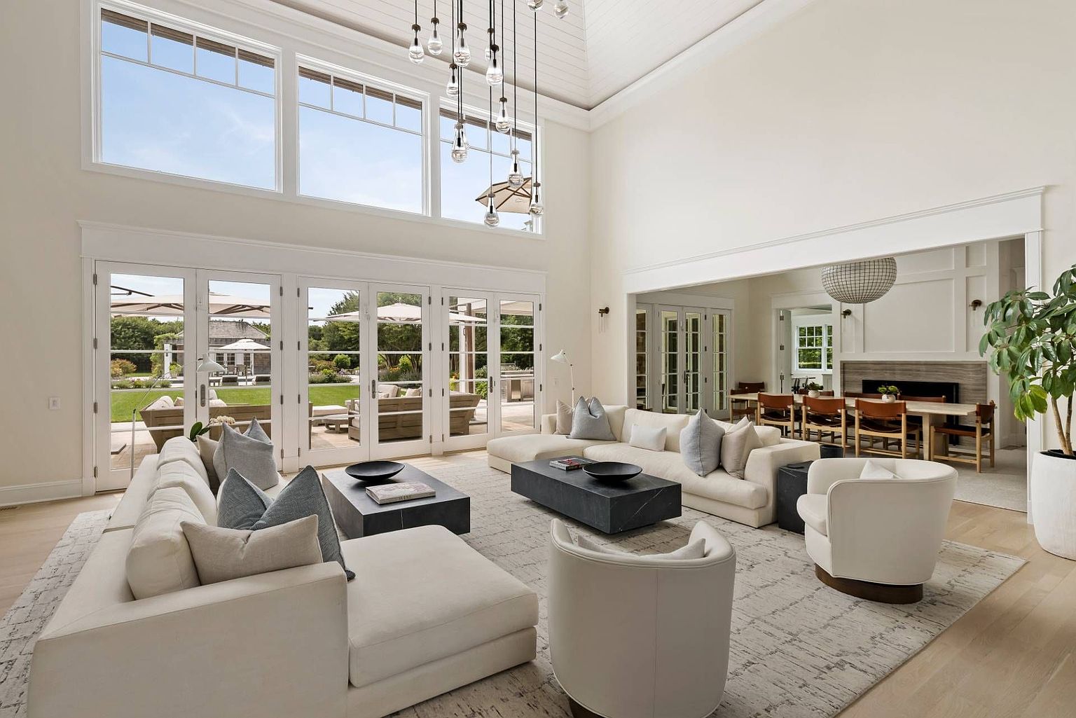 This expansive, light-filled living room features a double-height ceiling with a dramatic multi-pendant light fixture and large windows that provide a seamless connection to the outdoor patio. The space is anchored by two large, neutral-toned sectional sofas and two curved accent chairs arranged around dark stone coffee tables on a textured area rug. Beyond the main seating area, a wide opening leads into a formal dining room, creating an open-concept flow that emphasizes the home's airy and sophisticated design.
