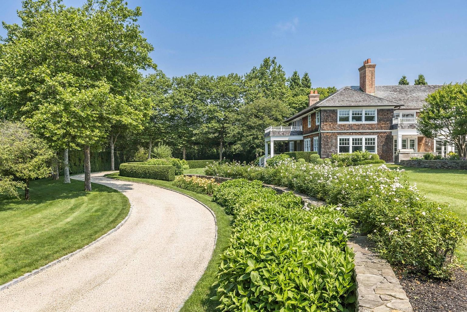 This picturesque exterior view showcases a winding gravel driveway leading toward a stately shingle-style residence surrounded by lush, manicured landscaping. A stone retaining wall borders a vibrant garden bed filled with blooming white roses and dense greenery, creating a classic, elegant curb appeal. The scene is captured from a ground-level perspective, emphasizing the expansive lawn and the mature trees that frame the property.