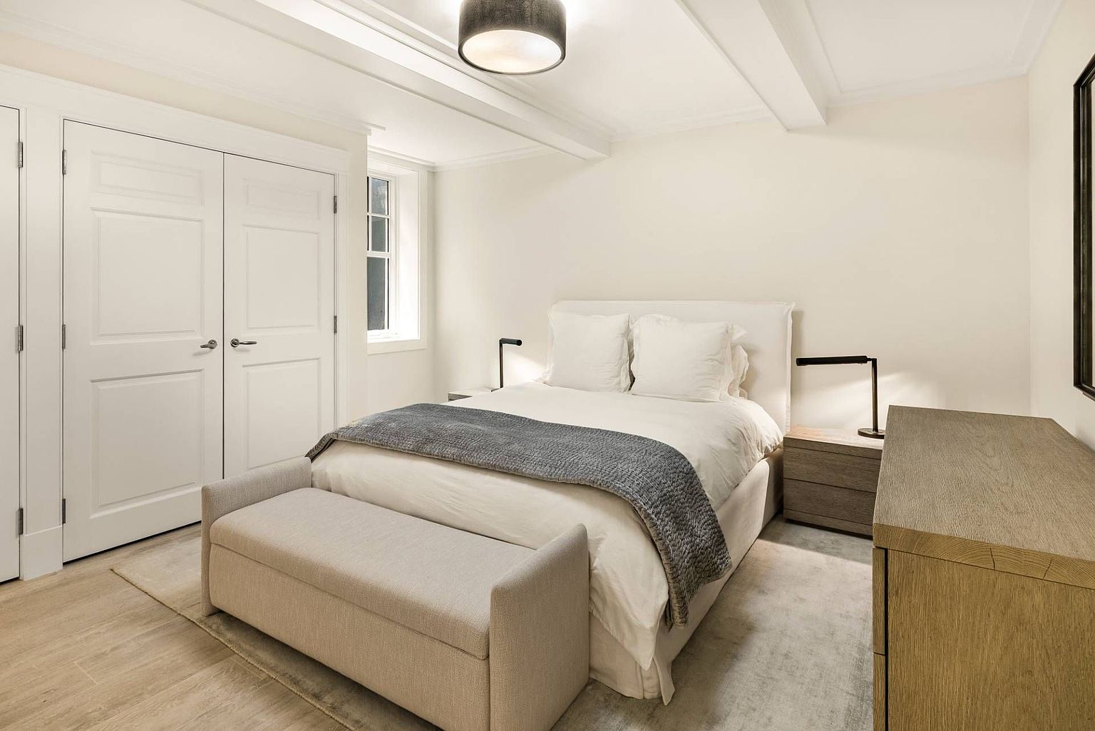 This bright and neutral bedroom features a queen-sized bed dressed in crisp white linens with a textured grey throw blanket. A matching upholstered bench sits at the foot of the bed, while a light wood dresser and a minimalist bedside lamp complete the clean, contemporary aesthetic. The room is accented by white paneled closet doors and a single window, creating a calm and inviting atmosphere.