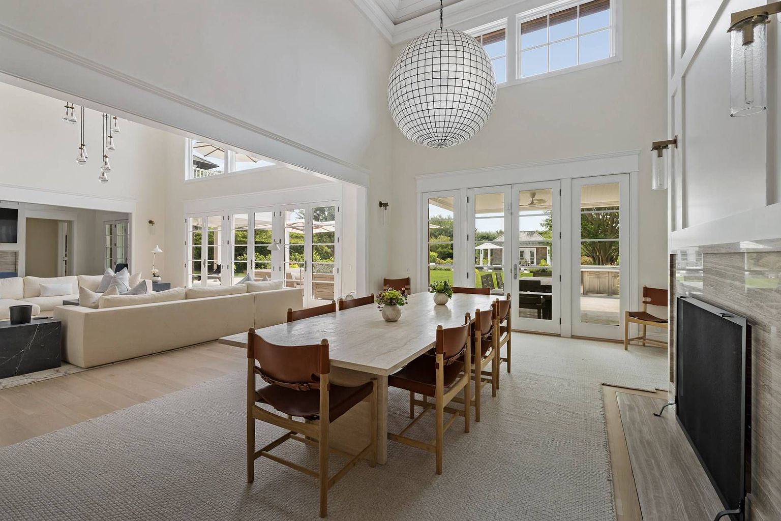This spacious, open-concept dining area features a long, light-toned table surrounded by leather-upholstered chairs, positioned beneath a large, spherical grid-patterned pendant light. The room flows seamlessly into an adjacent living area, with tall glass doors providing views of the outdoor landscape and abundant natural light. A stone-clad chimney breast anchors the side of the room, contributing to the airy, sophisticated, and modern aesthetic of the home.