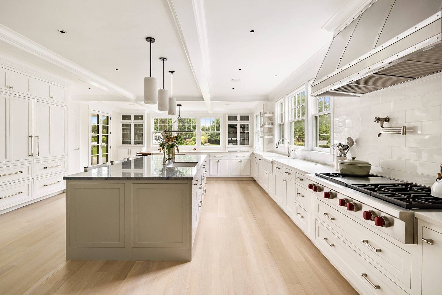 This bright and spacious kitchen features white cabinetry, a large central island with a stone countertop, and light wood flooring. A professional-grade range with a prominent stainless steel hood serves as a focal point, complemented by subway tile backsplash and pendant lighting. The perspective is a wide-angle shot from the center of the room, showcasing an open layout with ample natural light streaming through multiple windows.