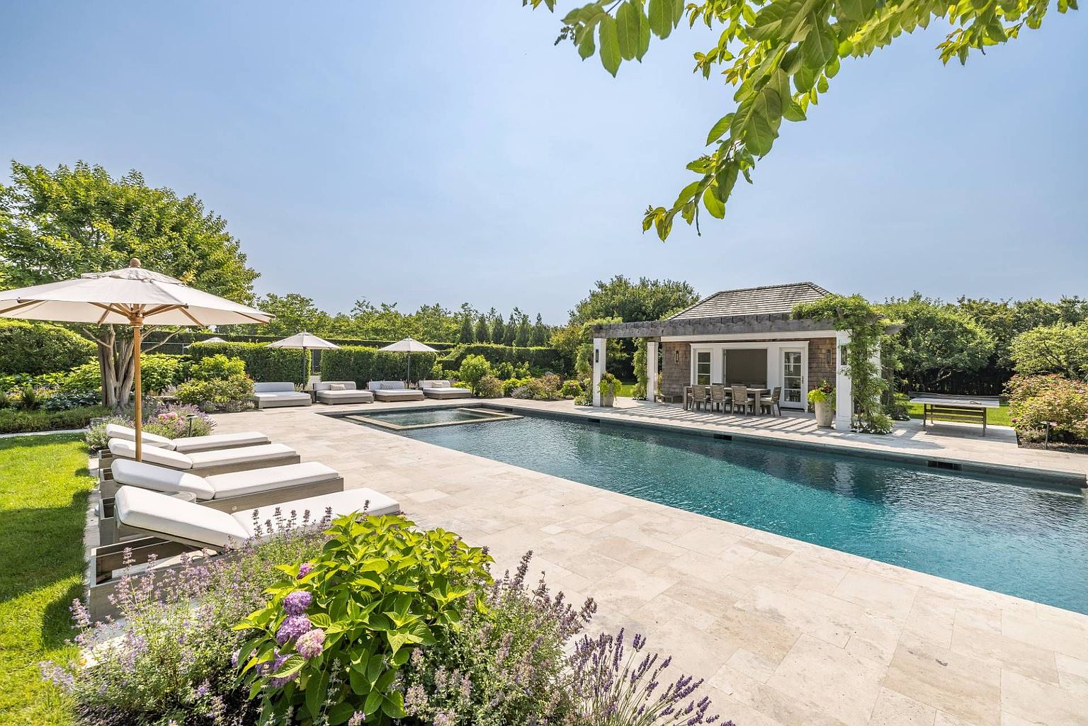 This expansive outdoor living space features a long, rectangular swimming pool with an integrated spa, surrounded by light-toned stone decking. Multiple lounge chairs with umbrellas are arranged along the pool edge, while a covered cabana with an outdoor dining set provides a shaded retreat. The scene is framed by lush green hedges, mature trees, and vibrant lavender plantings in the foreground, creating a serene and luxurious backyard atmosphere.