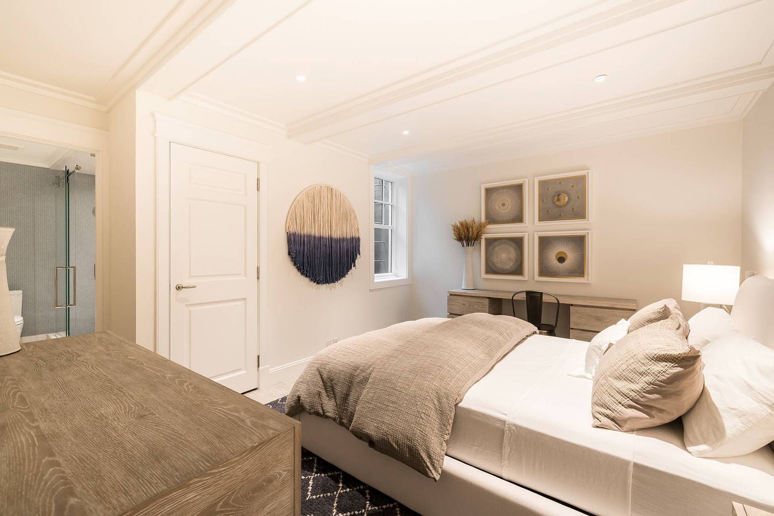 This bright and serene bedroom features a neutral color palette with a plush bed dressed in soft linens and a textured throw. A light wood dresser sits against the wall, accented by a set of four framed art pieces and a decorative wall hanging, while a glimpse into an adjacent bathroom reveals a modern glass-enclosed shower. The room is bathed in soft, warm lighting, creating a tranquil and inviting atmosphere.