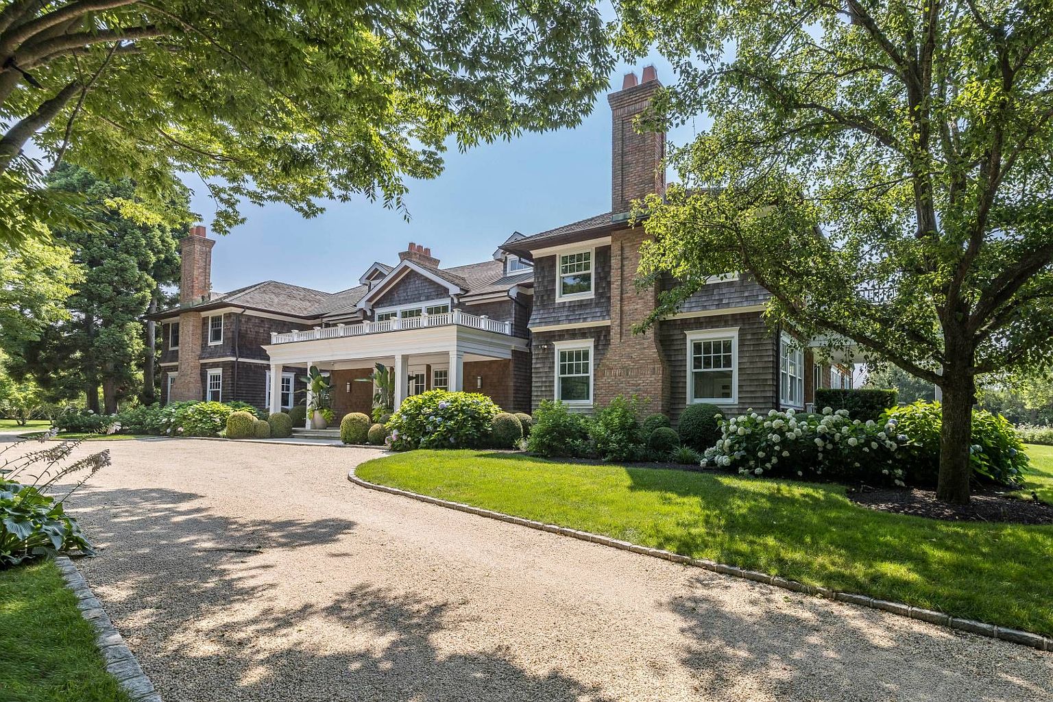 This grand, shingle-style estate features a classic architectural design with a prominent brick chimney and a welcoming covered portico entrance. The home is surrounded by lush, mature trees and meticulously landscaped gardens, with a sweeping gravel driveway leading up to the front facade. The perspective captures the stately presence of the residence, emphasizing its expansive scale and elegant, traditional curb appeal.