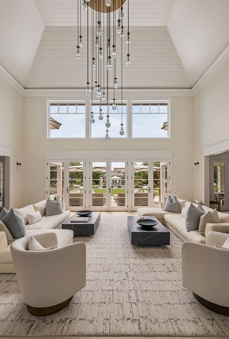 This expansive, symmetrical living room features a soaring vaulted ceiling with shiplap detailing and a dramatic, multi-pendant chandelier as the central focal point. The space is anchored by two large, light-colored sectionals facing each other across dark stone coffee tables, all set upon a textured area rug. Large windows and glass doors provide an abundance of natural light and a clear view of the manicured backyard, creating an airy and sophisticated atmosphere.
