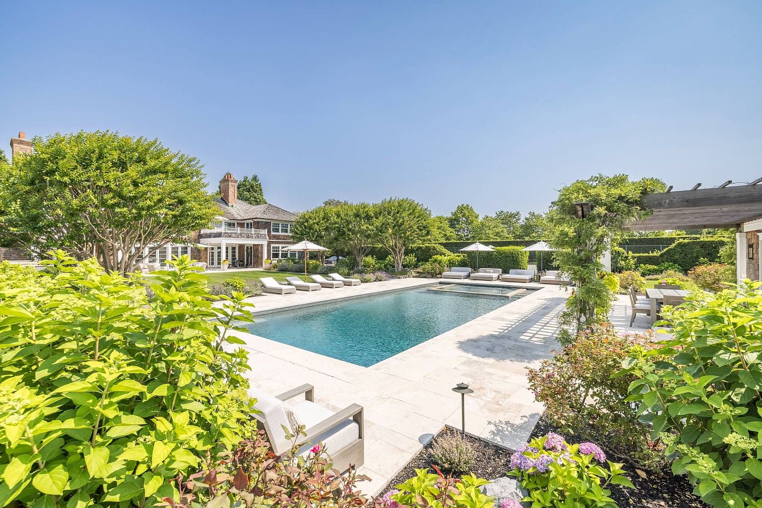 This expansive backyard features a rectangular swimming pool with an integrated spa, surrounded by light-toned stone decking and multiple lounge chairs. The scene is framed by lush green trees, vibrant hydrangea bushes, and a wooden pergola covering a dining area, creating a serene and private outdoor retreat. In the background, a classic shingle-style residence with a multi-gabled roofline completes the elegant, resort-like atmosphere.